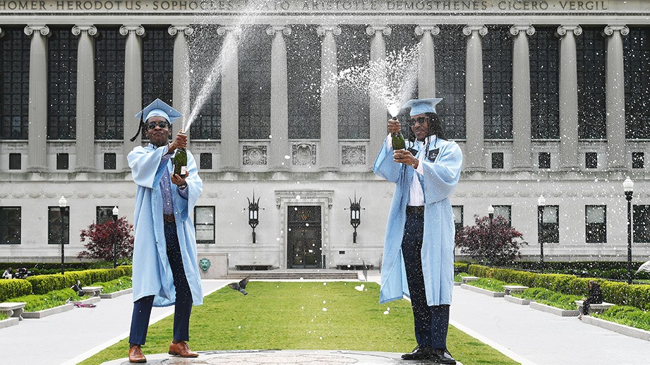 Photo by Eileen Barroso Two Columbia graduates in regalia pop champagne in front of Butler Library.