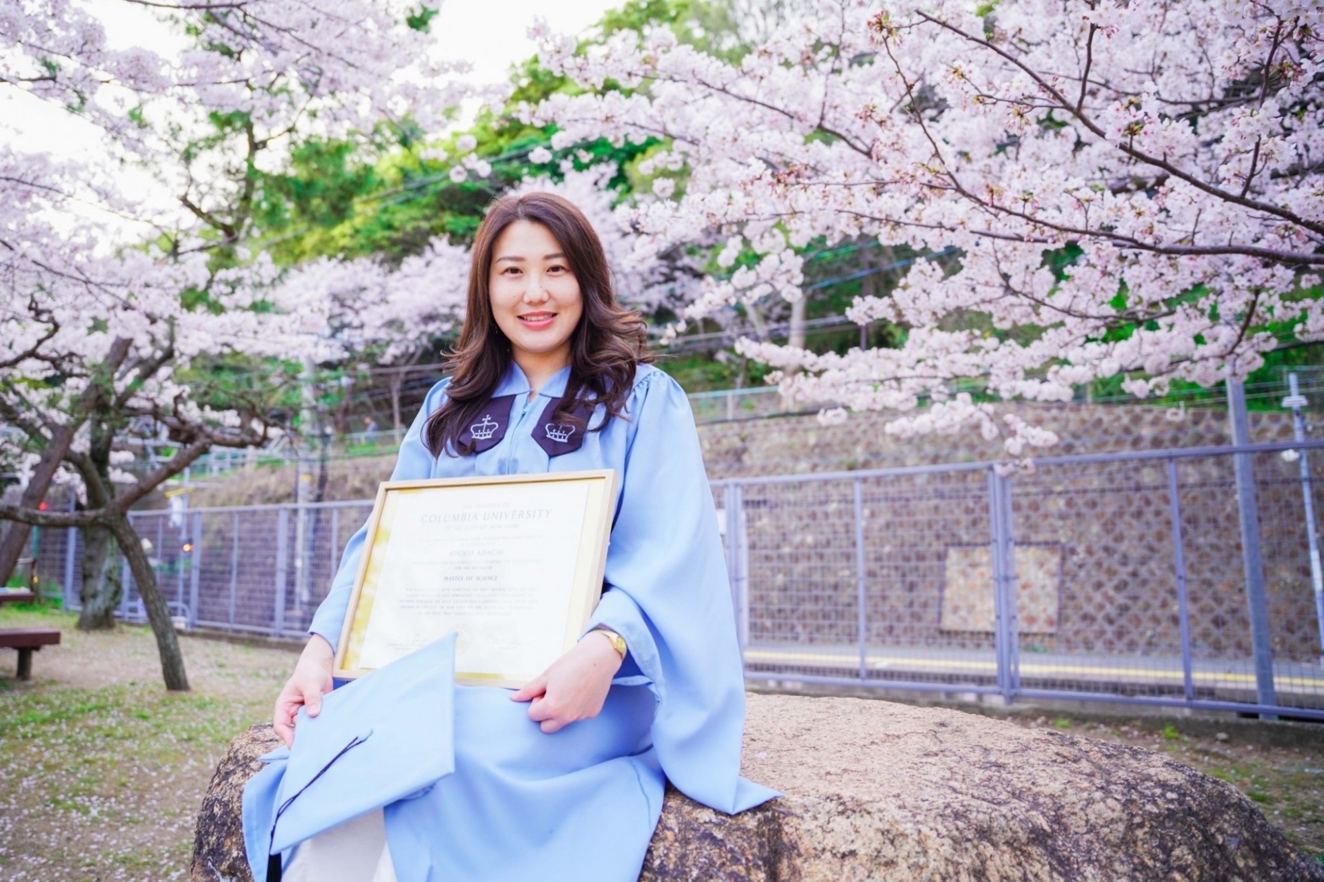 A Columbia grad in gown with diploma in Japan surrounded by cherry blossom trees. 