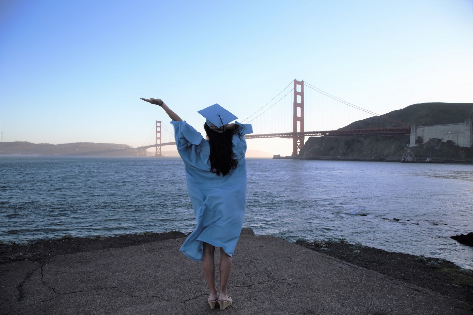 A Columbia graduate in cap and gown throws their hands up in front of the Golden Gate Bridge. 