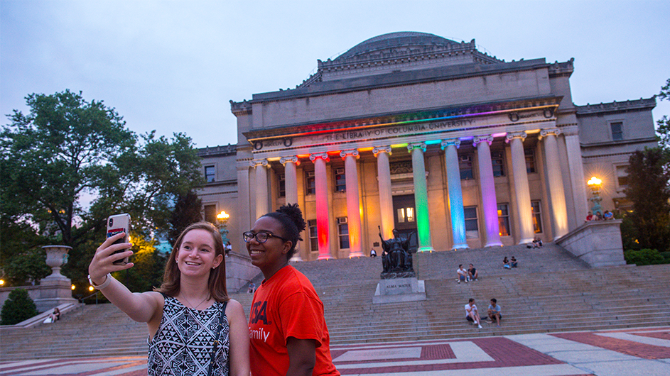 Two girls take a selfie in front of the Low Library columns, which are lit up in rainbow colors.