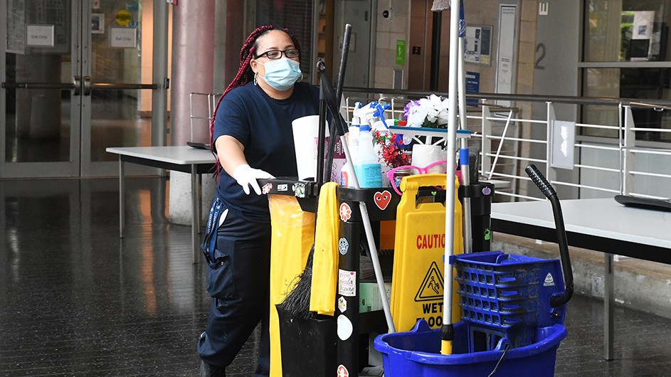 A custodion in braids and a face mask pushes a cart of cleaning supplies on Columbia's campus.