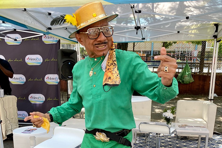 A man in a green suit and golden hat dances in front of a booth. 