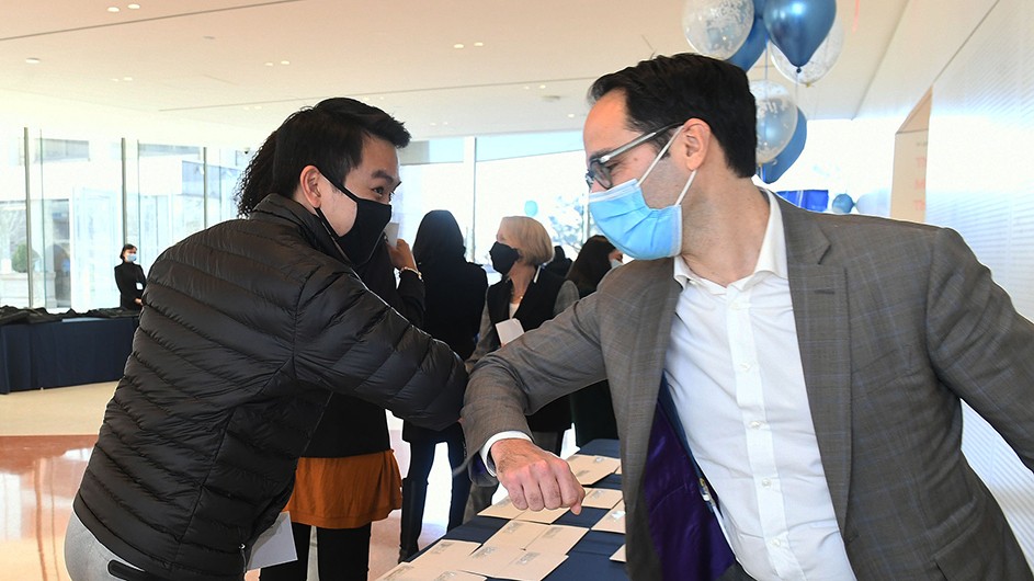 Two people with masks on bump elbows over Match Day envelopes.
