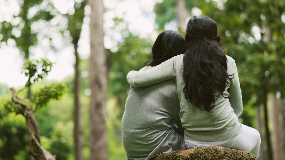 The backs of two individuals with long dark hair sitting next to one another in nature. One has their arm around the other. 