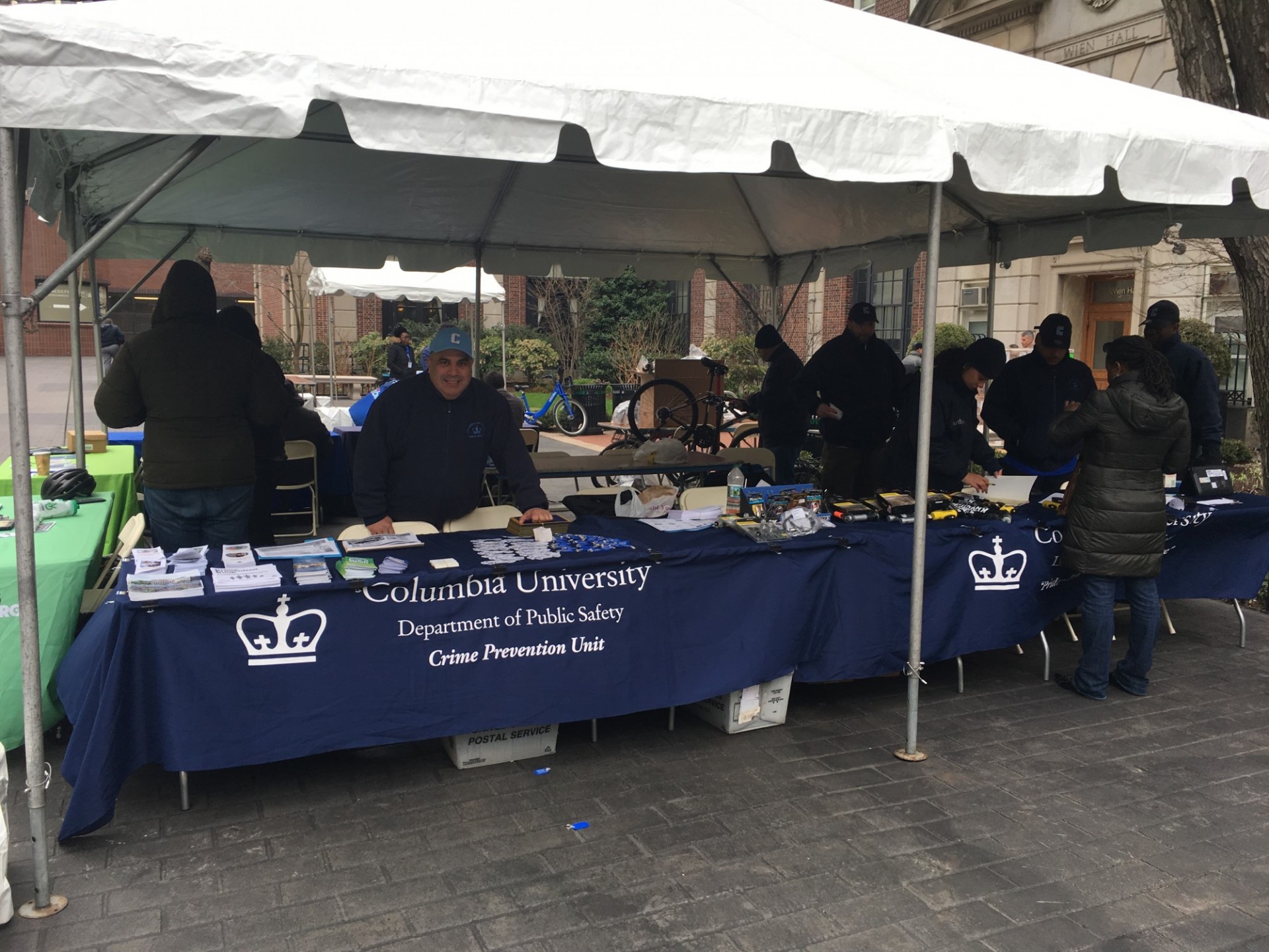 A tent outdoors shelters a group of people at a table. The table has a sign that reads Columbia University Public Safety.