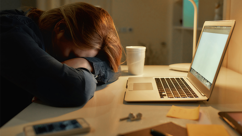 A woman collapses her head on her arms in front of a laptop with a coffee cup beside her.