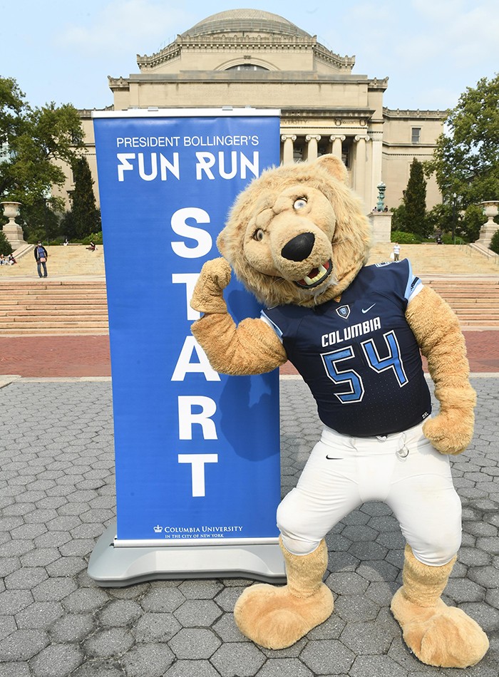 A lion mascot stands in front of a President Bollinger Fun Run sign. 
