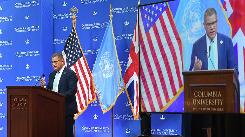 COP26 President Alok Sharma in navy blue suit speaks at a podium with Columbia signage and banner in front of UN, American, and British flags on stage at Columbia University Low Library.