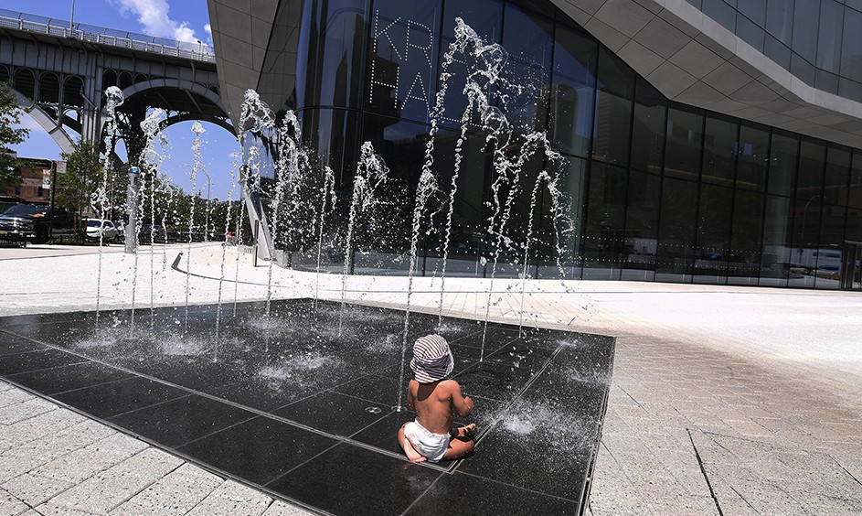 A young child in diapers and hat playing in a water sprinkler next to Kravis Hall on a hot sunny day at Columbia Manhattanville in New York City