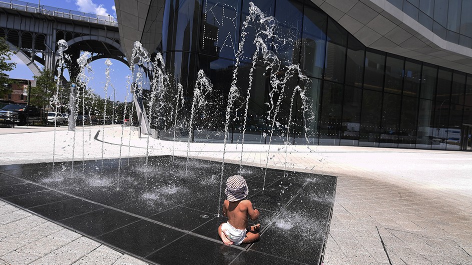 A young child in diapers and hat playing in a water sprinkler next to Kravis Hall on a hot sunny day at Columbia Manhattanville in New York City