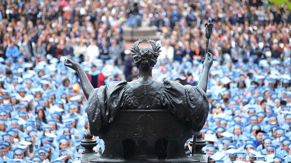 Alma Mater statue overlooks a crowd of blue caps and gowns. 