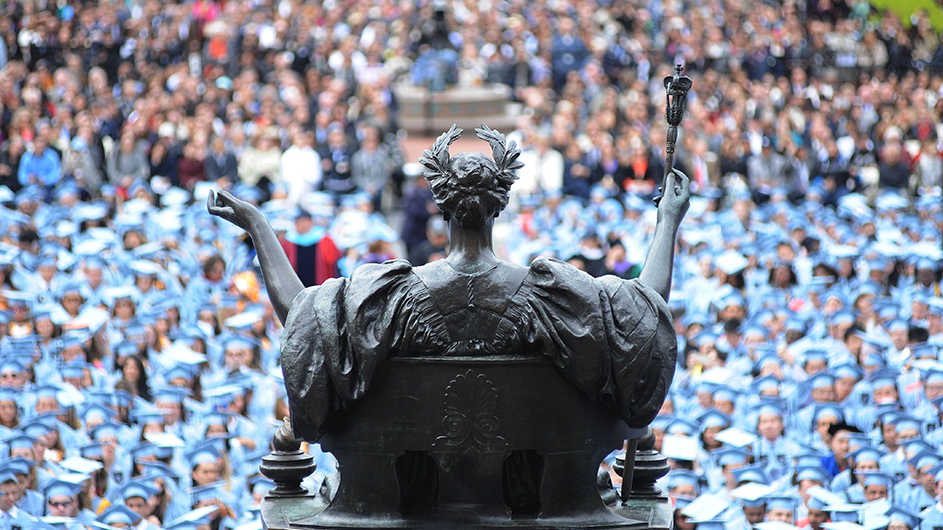 A view from behind the Alma Mater of a sea of blue caps and gowns. 