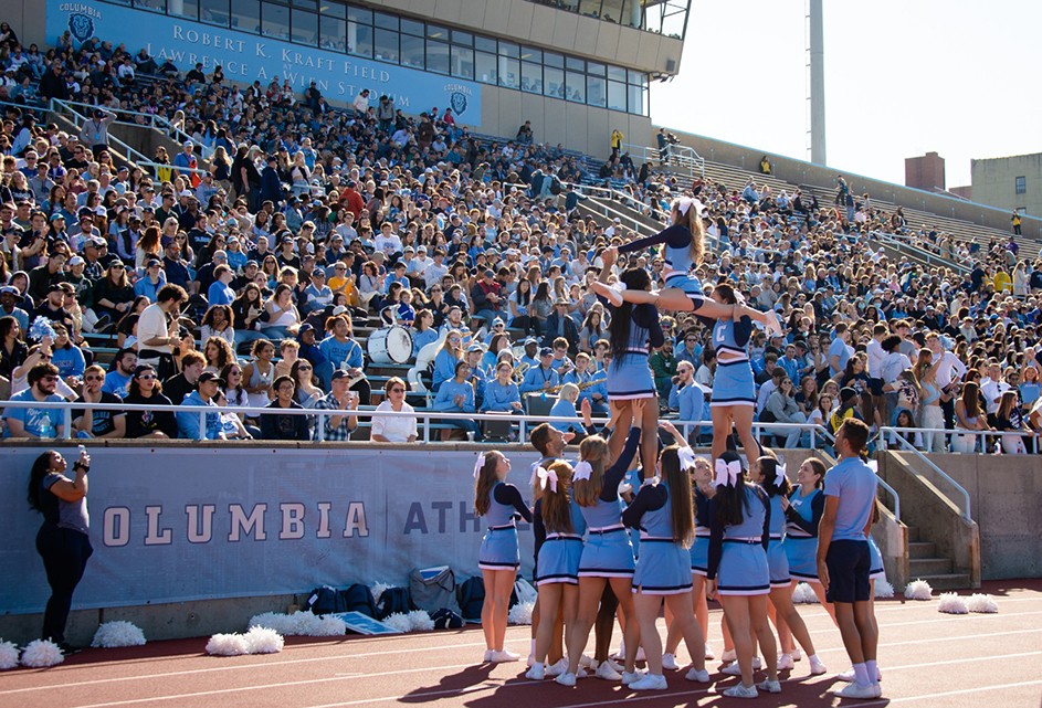 A dozen Columbia cheerleaders in light blue uniforms cheer in a pyramid and splits formation in front of hundreds of spectators at Columbia's Homecoming football game at Robert K. Kraft Field at Baker Athletics Complex in upper Manhattan.