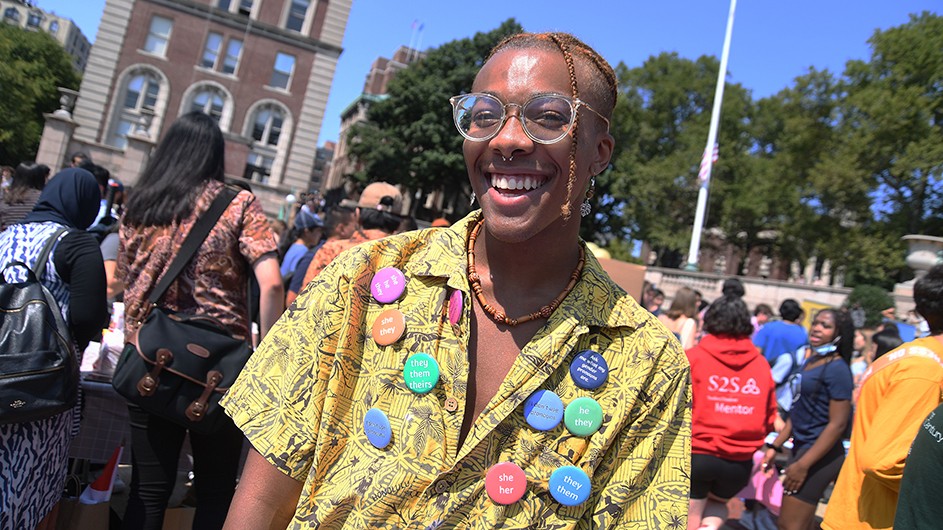 A student in bright yellow shirt with 10 colorful buttons of different pronouns smiles at the activities fair on a sunny day at Columbia University.