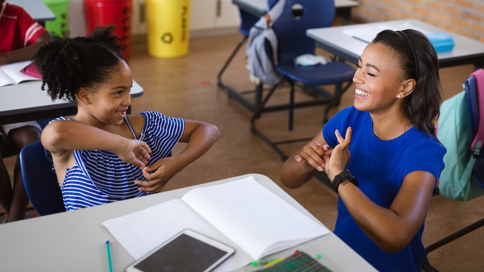 A young girl and her teacher smiling and speaking in sign language at a classroom table