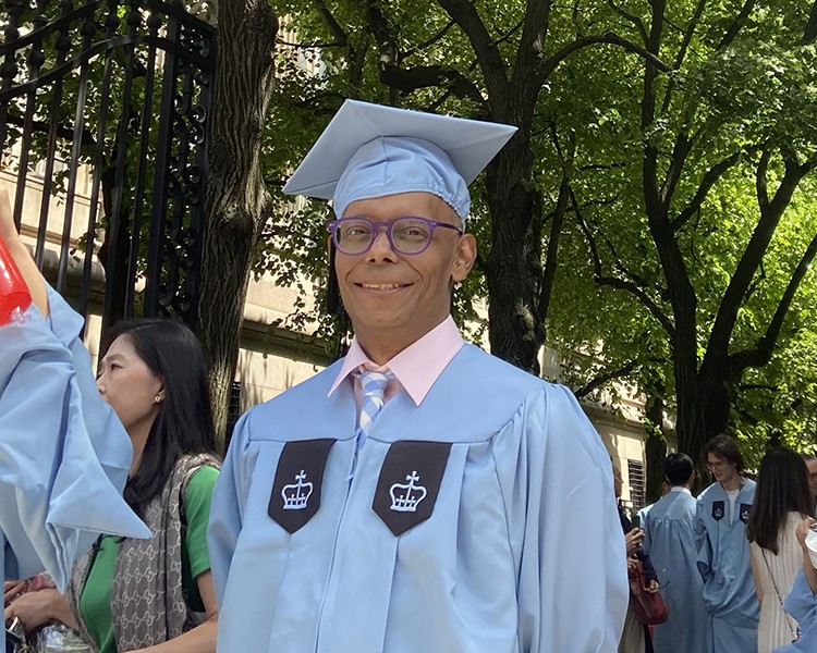 Freddy Claudio posing in graduation cap and gown at Columbia University's College Walk gates.