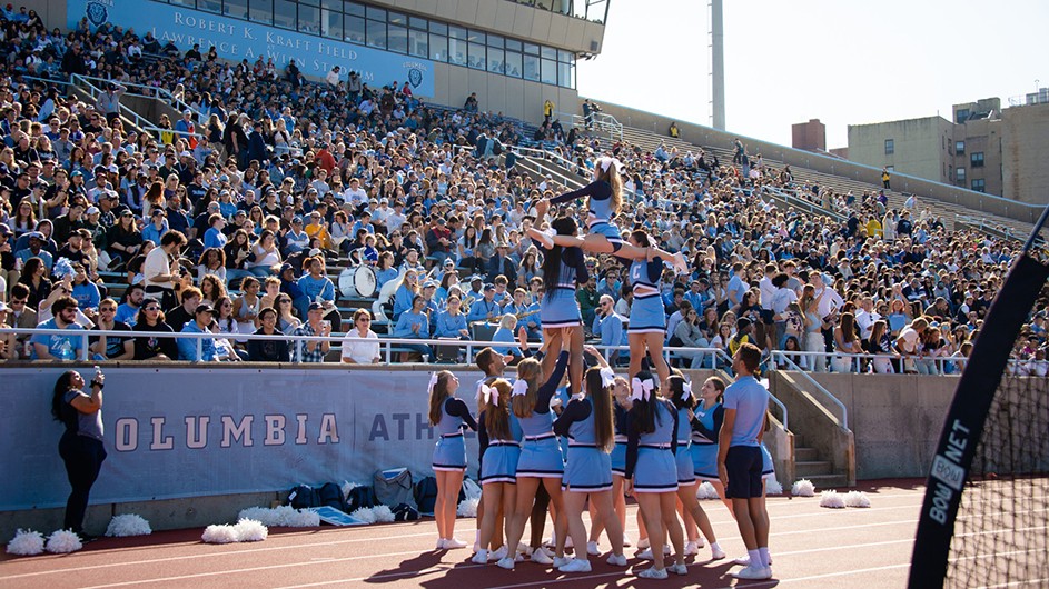 A dozen Columbia cheerleaders in light blue uniforms cheer in a pyramid and splits formation in front of hundreds of spectators at Columbia's Homecoming football game at Robert K. Kraft Field at Baker Athletics Complex in upper Manhattan.