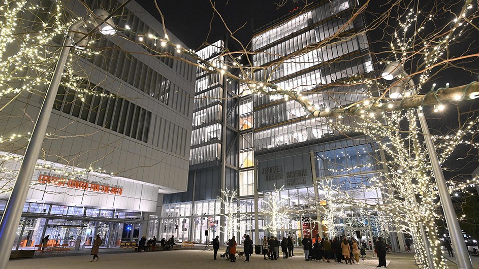 A crowd gathers on the lighted holiday tree-lined plaza in front of Jerome L. Greene Science Center and Lenfest Center for the Arts at Columbia Manhattanville at night.