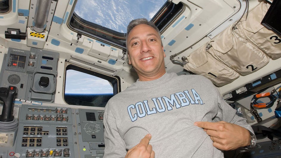 Astronaut Mike Massimino in space smiles while pointing to his Columbia shirt.