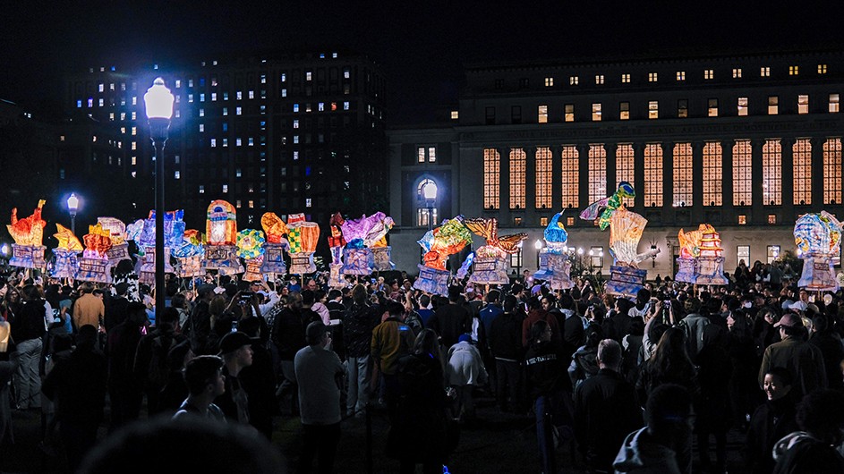 Dozens of people carrying colorful illuminated lanterns alongside hundreds of spectators at night in front of Butler Library at Columbia University. 