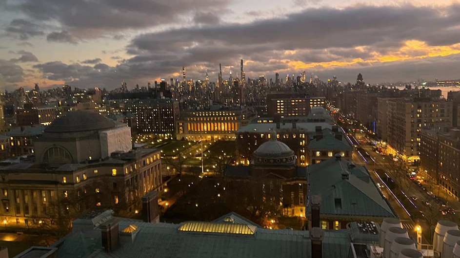 Columbia University's illuminated Morningside campus with New York City's skyline in the distance with a partly-cloudy sky at dusk.