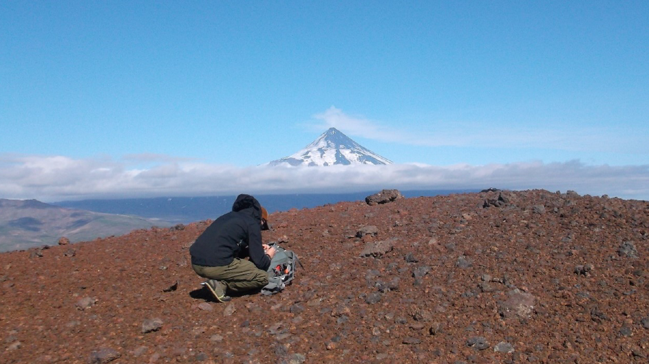 collecting volcanic ash from the Aleutian Islands’ Fisher Caldera with Shishaldin volcano in the distance. (Diana Roman/Carnegie Institution for Science)