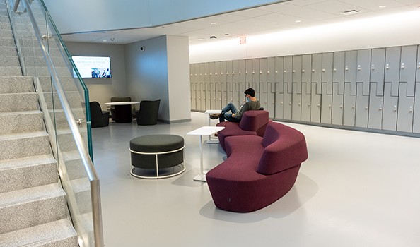 A student sitting on a purple sofa in front of a long hallway of silver lockers at Columbia Business School