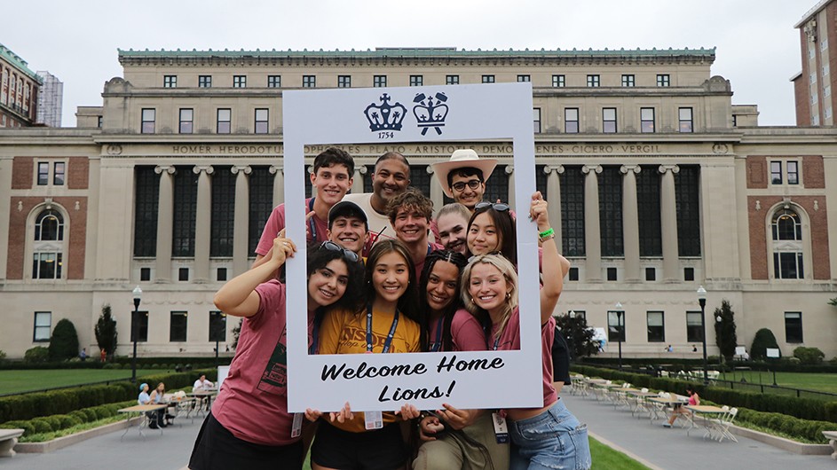 Eleven students grouped together smile through a large Welcome Home Lions! picture frame on the Sundial in front of Butler Library at Columbia University on a summer day.