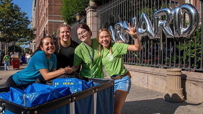 Four women pose in front of Barnard balloons with a moving bin. 