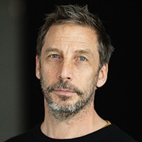 Photo: Lisa Whiting. A person with short hair and a beard, against a black background.