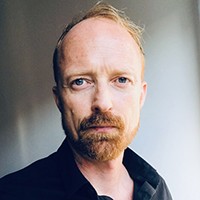 A man with short light hair in a beard and mustache, in a black top.