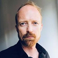 A man with short light hair in a beard and mustache, in a black top. 