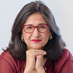 Minouche Shafik, a woman with long dark hair, in red glasses and a red top. 