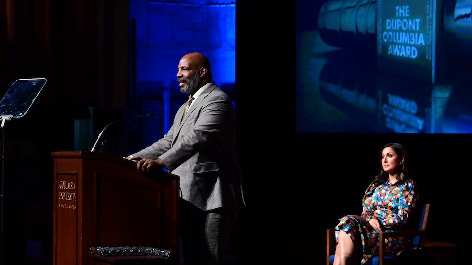 Jelani Cobb speaking at a podium at the 2023 DuPont-Columbia Awards. A women in a floral dress is seated in the background.