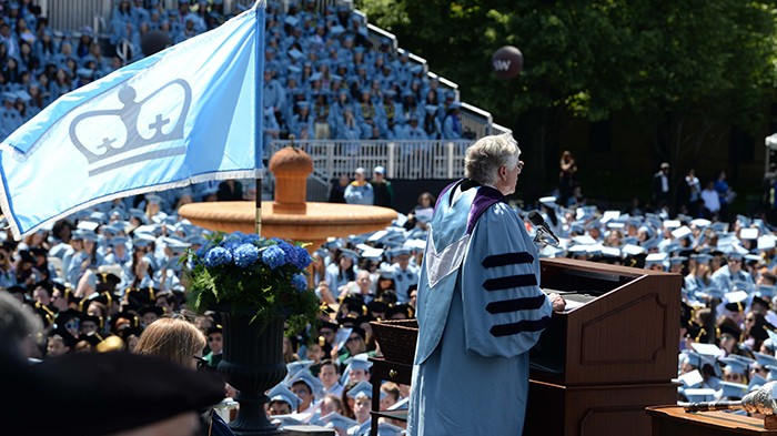 2019 photo by Eileen Barroso. President Bollinger addresses the commencement crowd as a flag with Columbia's crown flies behind him.
