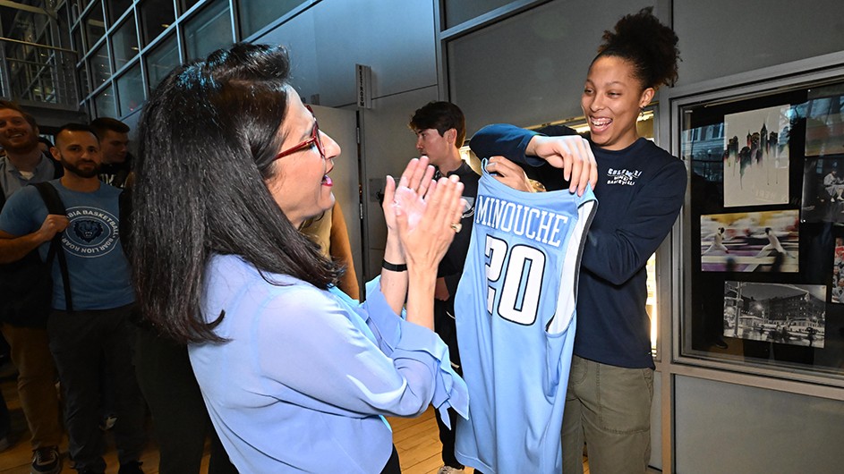 Columbia student-athlete Kaitlyn Davis smiles while holding up a light blue basketball jersey with the name "Minouche" for Columbia's next president Minouche Shafik who is smiling and clapping her hands in Lerner Hall at Columbia University.