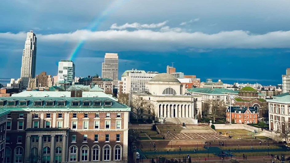 A colorful rainbow arches in a partly cloudy blue sky from the Northwest Corner Building and Pulitzer Hall over Low Library and Uris Hall at Columbia University.