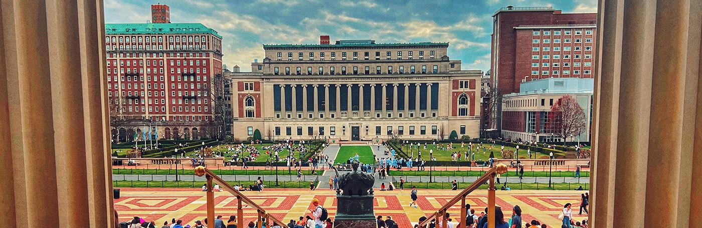 A view flanked by two Low Library center columns toward Alma Mater statue and Butler Library with dozens of students sitting on Low Library steps and South Lawn fields on a cloudy day at Columbia University.