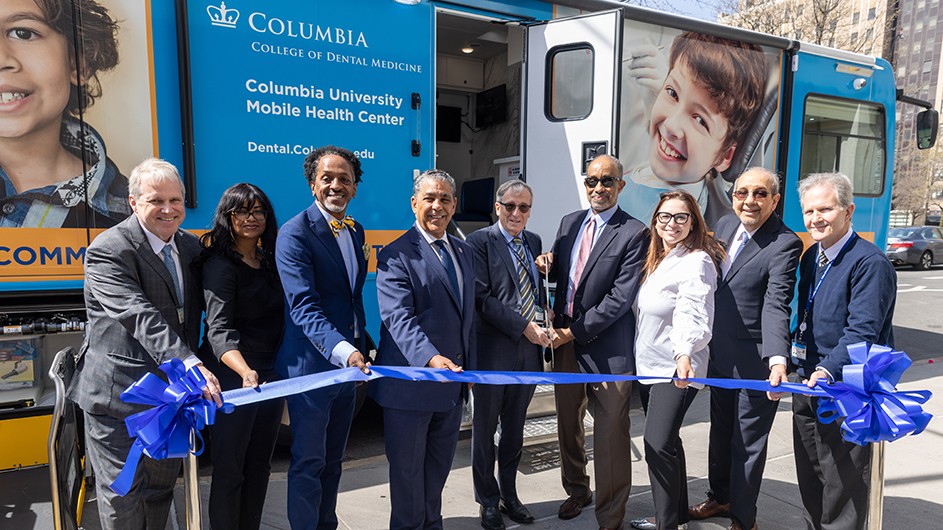 Photo by Chris Balmer/Columbia University Irving Medical Center. From left: Joseph Harney, vice dean of finance and administration; Amy Herbert, DDS, assistant professor of dental medicine; Al Taylor, New York State assemblymember; Adriano Espaillat, U.S. Congressional representative; Christian Stohler, DMD, dean of the College of Dental Medicine; Dennis Johnson, Mother Cabrini Health Foundation; Biana Roykh, DDS, associate professor of dental medicine and senior associate dean for clinical affairs; Walid Michelen, MD, ArchCare; Marc Michalowicz, DDS, director for management of vulnerable populations, College of Dental Medicine. Nine people in suits and work attire smile and pose to cut the celebratory opening ribbon in front of a blue Columbia University medical and dental van in Upper Manhattan.