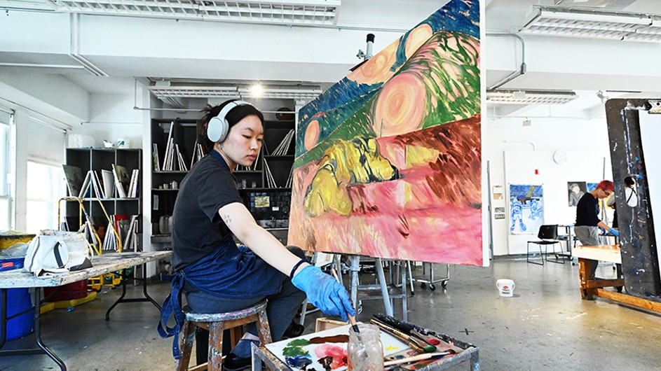 A student in headphones paints a bright-colored canvas in a sunny room in Dodge Hall at Columbia University.