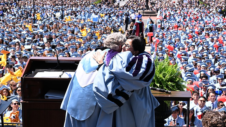 President Lee Bollinger and President-Designate Minouche Shafik, both dressed in Columbia regalia, embrace on stage with hundreds of graduates looking on at Columbia Commencement on a sunny day.