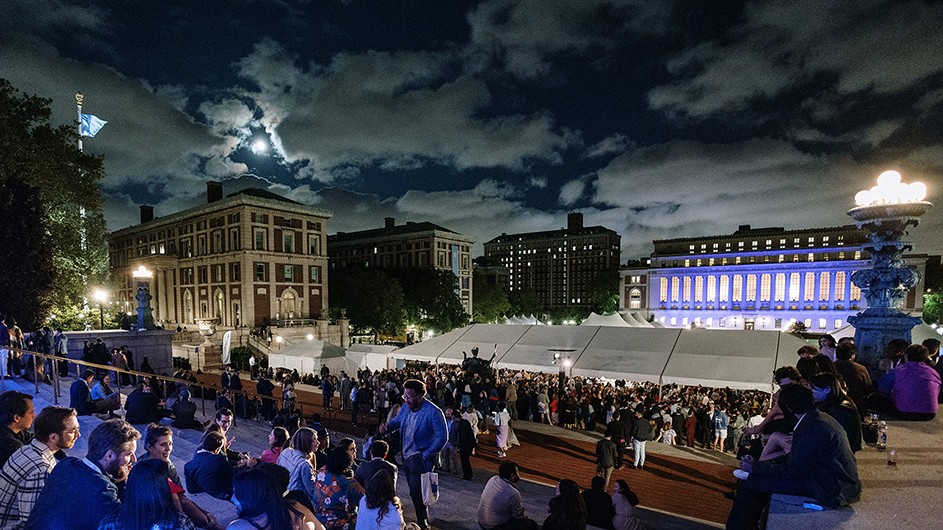 Hundreds of Columbia alumni celebrate their reunion on Low Library steps and under a huge party tent on Low Plaza with illuminated Butler Library in the background at Columbia University on a partly cloudy moonlit night.