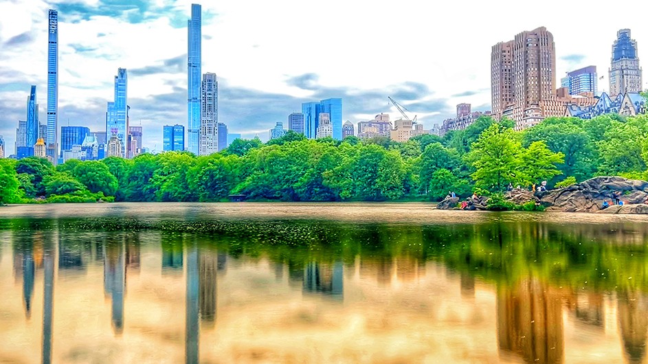 Central Park pond reflected with and lined by green trees and skyscrapers on a cloudy day in New York City.
