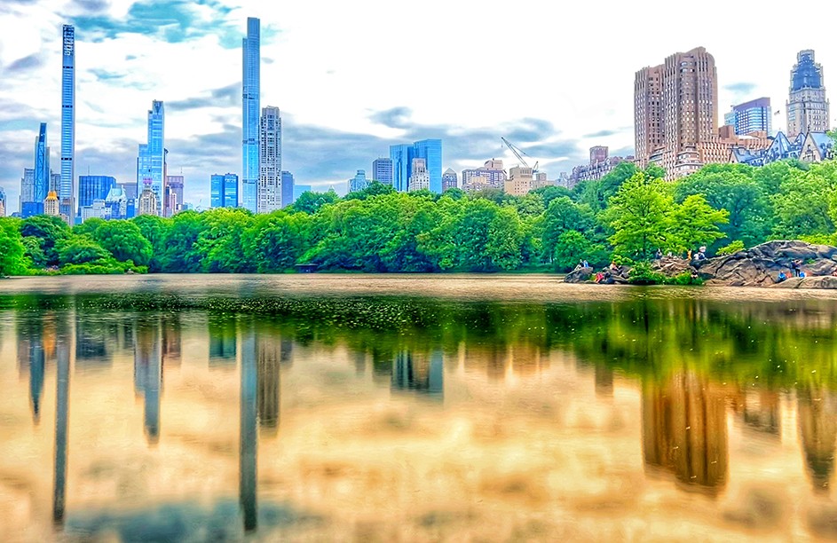 Central Park pond reflected with and lined by green trees and skyscrapers on a cloudy day in New York City.