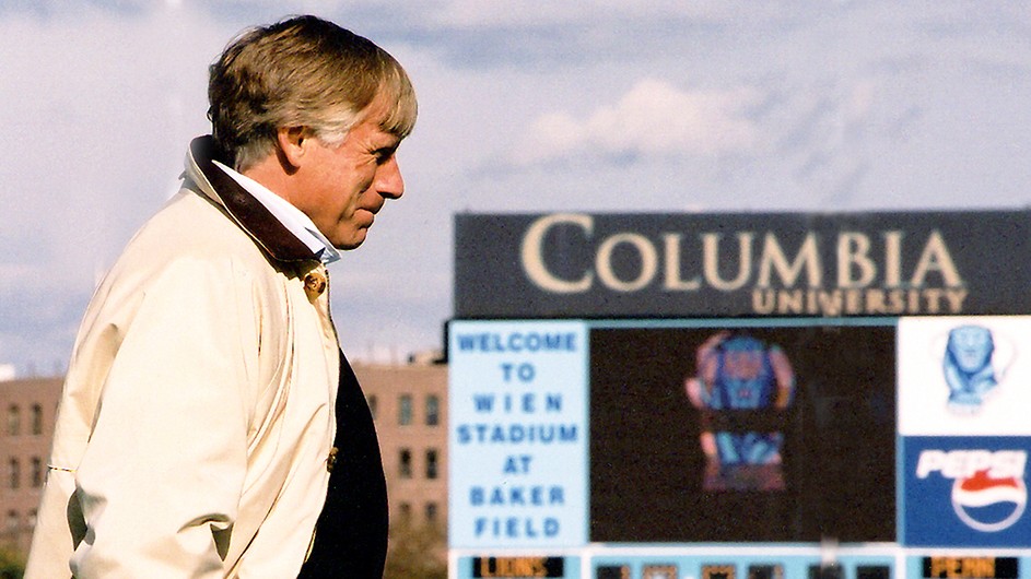 Columbia President Lee Bollinger in profile smiles with a Columbia University scoreboard in the background at Baker Athletics Complex on a partly cloudy day.