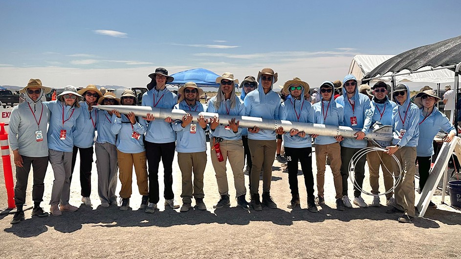 Fifteen Columbia University students in light blue shirts pose while holding a long hybrid rocket on a sunny day.