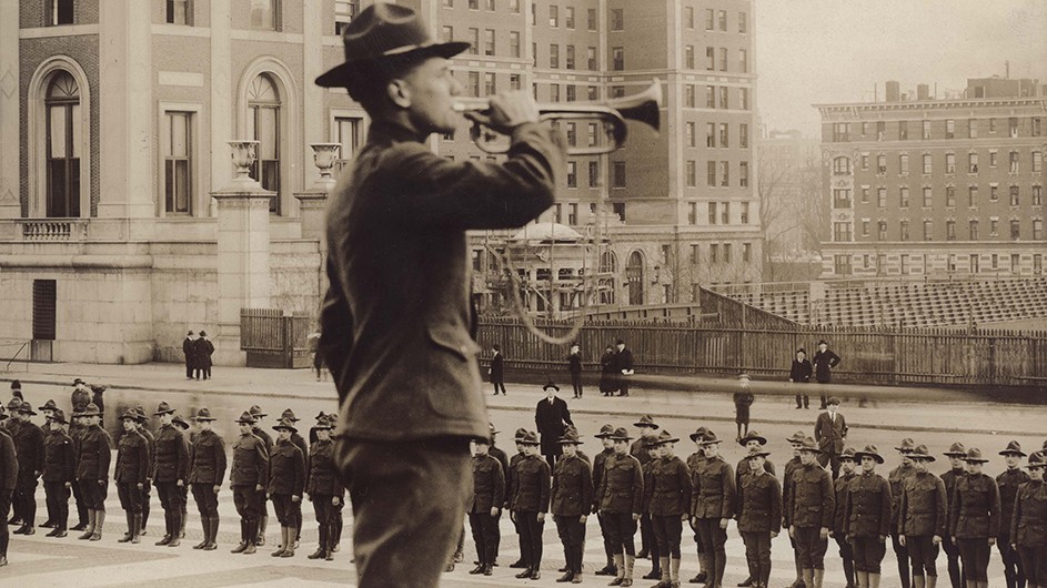 A student in 1918 wearing a WWI military uniform playing the bugle in front of dozens of uniformed students standing at attention on Low Plaza at Columbia University.  