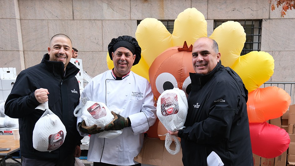 Three Columbia employees (Gregorio Matos, Chef Mike DeMartino, and Ricardo Morales) smile while holding up frozen turkeys in front of an inflated turkey balloon at a Thanksgiving turkey donation drive on 114th Street near Columbia University.