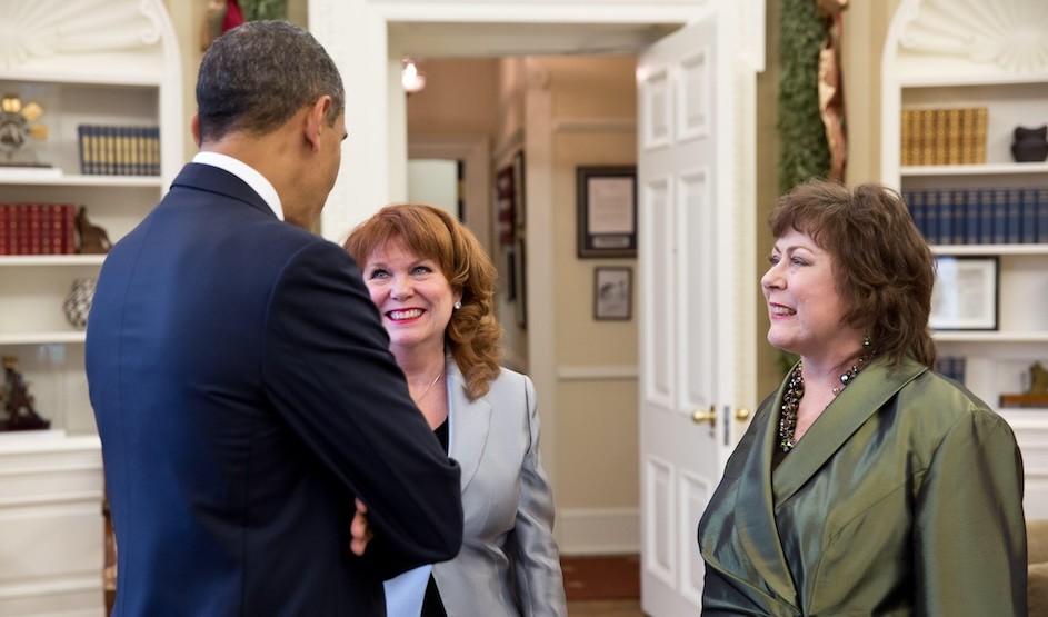 President Obama stands with his back to the camera in the foreground, engaged in conversation with two smiling women, who face the camera.