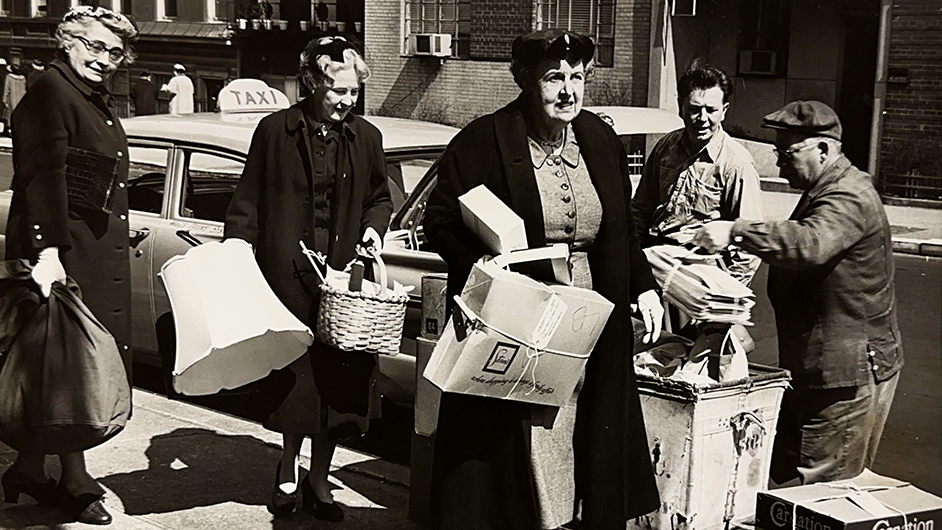 Black and white photo of women carrying lamps and boxes from a taxi. 